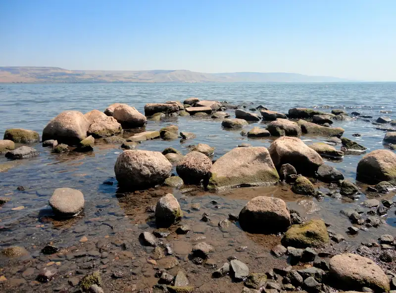 Peaceful rocky shore of a tranquil lake with clear blue sky and distant hills.